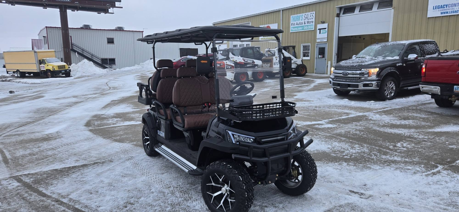 Black golf cart parked on a snowy lot near buildings and other vehicles.