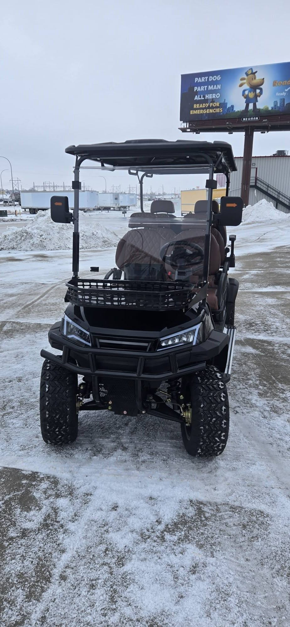 Black utility golf cart parked on a snowy lot under a blue sky, with a billboard in the background