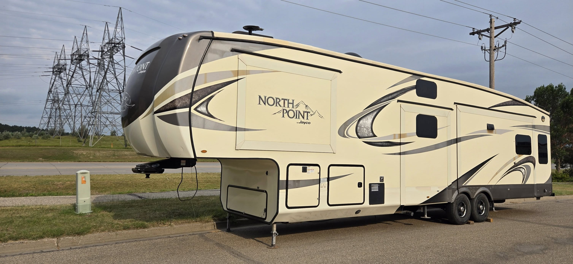 Beige and black fifth-wheel RV parked beside a road under a cloudy sky