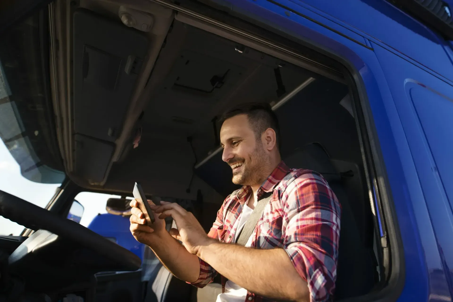 Homem sorrindo na cabine de um caminhão azul, olhando para o celular.