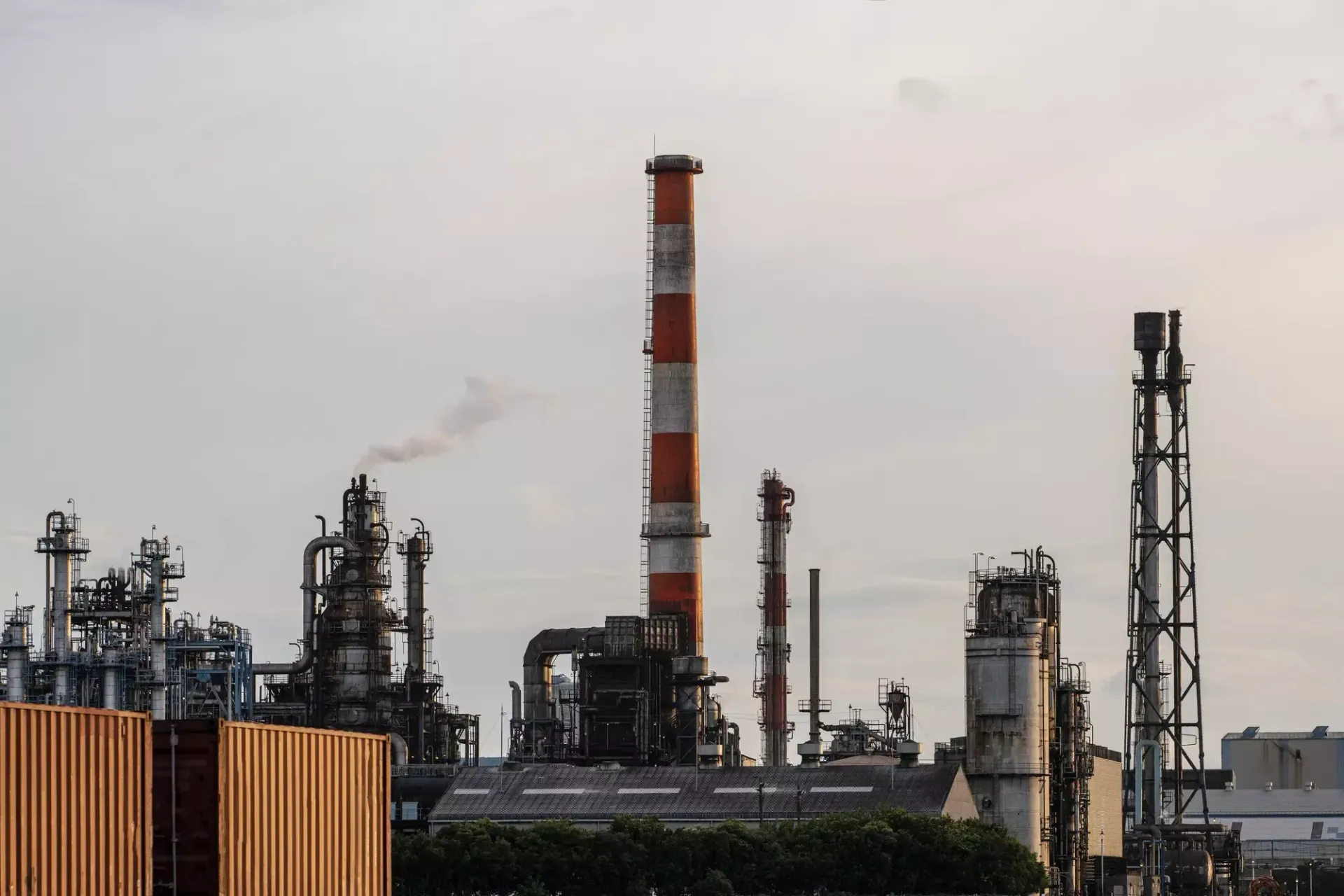 Refinaria industrial com chaminés altas e fumaça cinza contra um céu nublado.