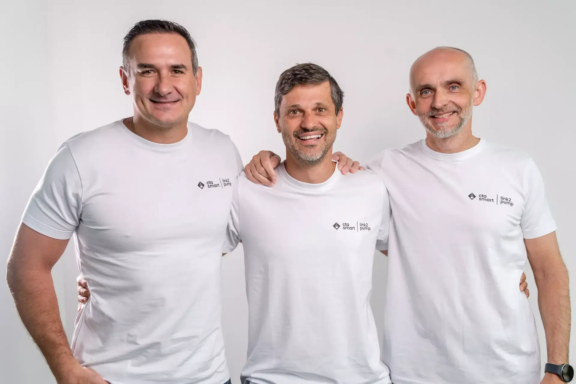 Três pessoas sorridentes vestindo camisetas brancas, em pé juntas contra um fundo claro.
