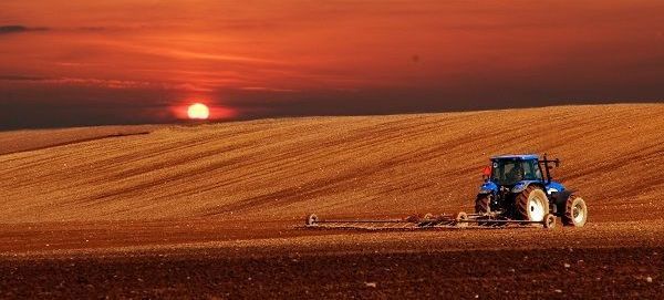 Trator azul arando um campo vermelho-acastanhado ao pôr do sol sob um céu alaranjado brilhante.
