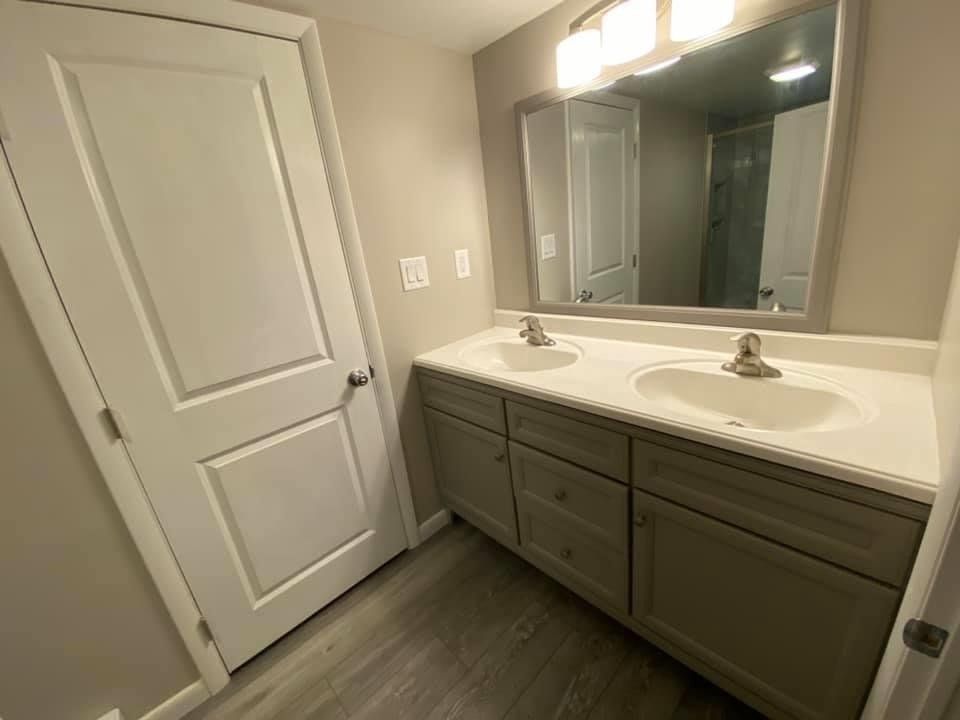 Bathroom with double-sink vanity, large mirror, gray cabinets, and a closed door.