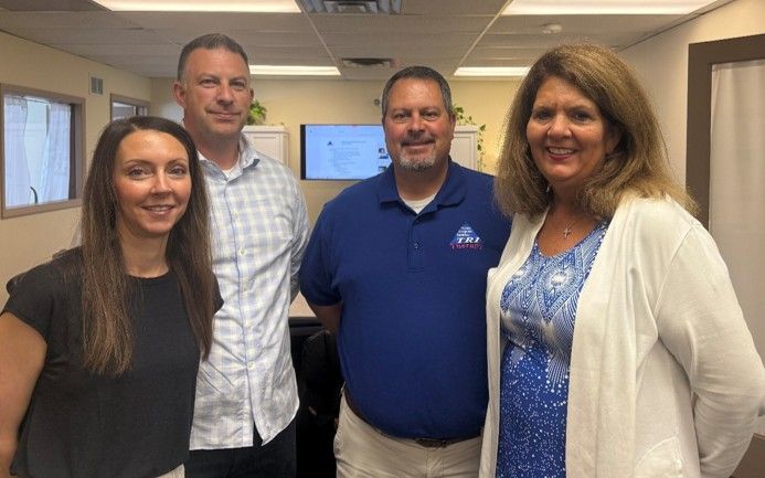 Four coworkers smiling in an office hallway, standing together under fluorescent lights.