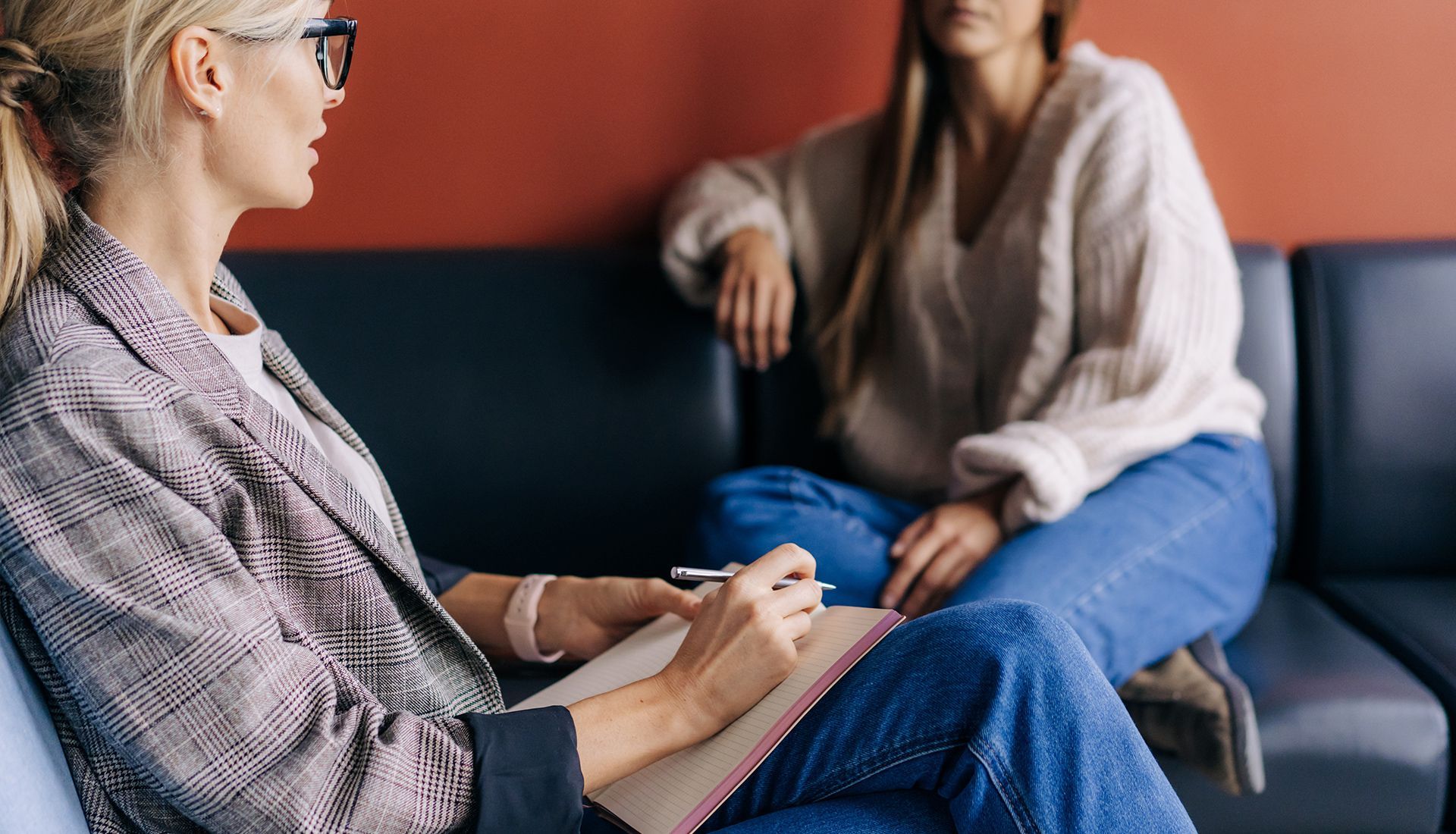 A therapist takes notes while listening to a client seated on a black couch in a counseling session.