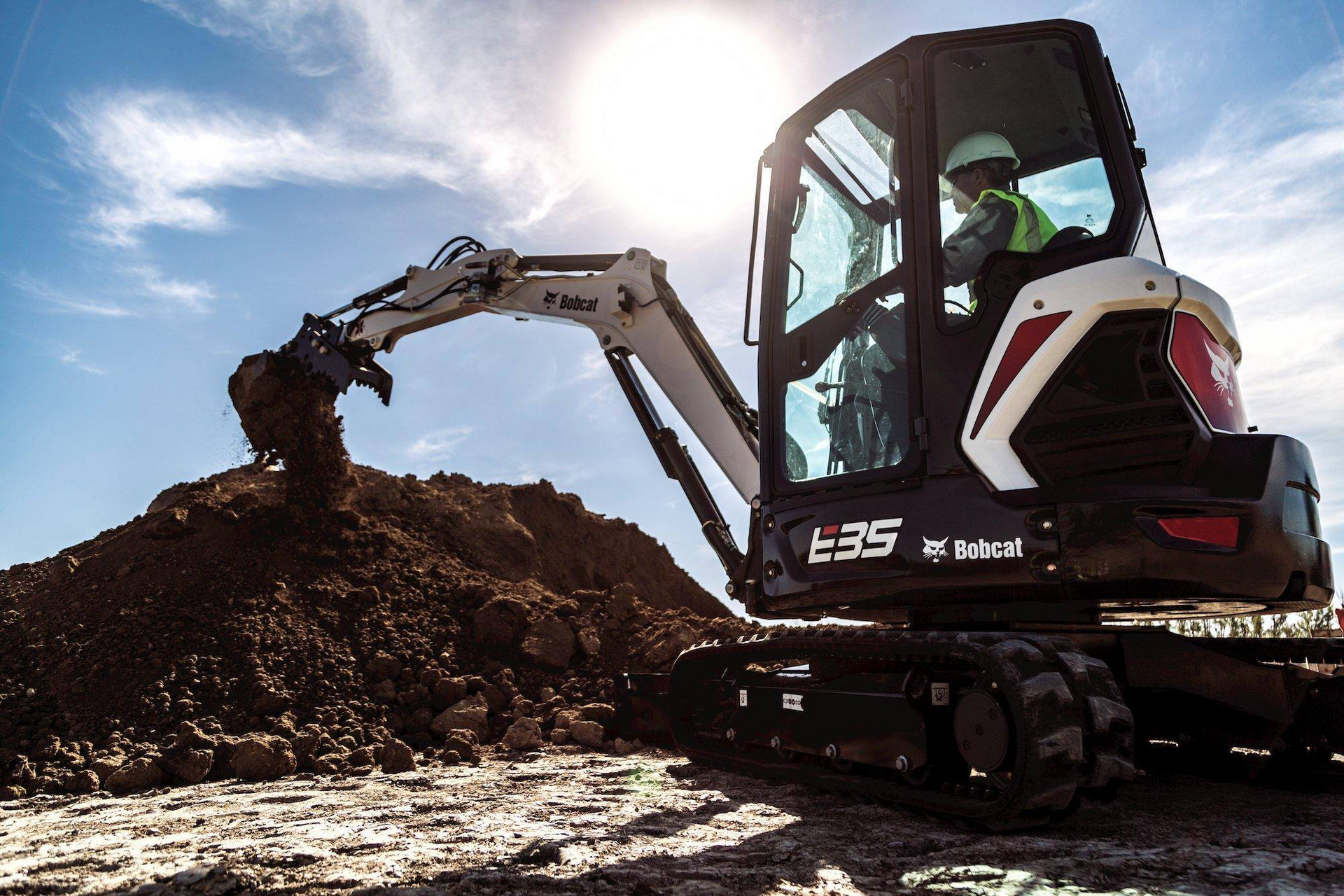 Bobcat excavator digging dirt pile; operator in cab.