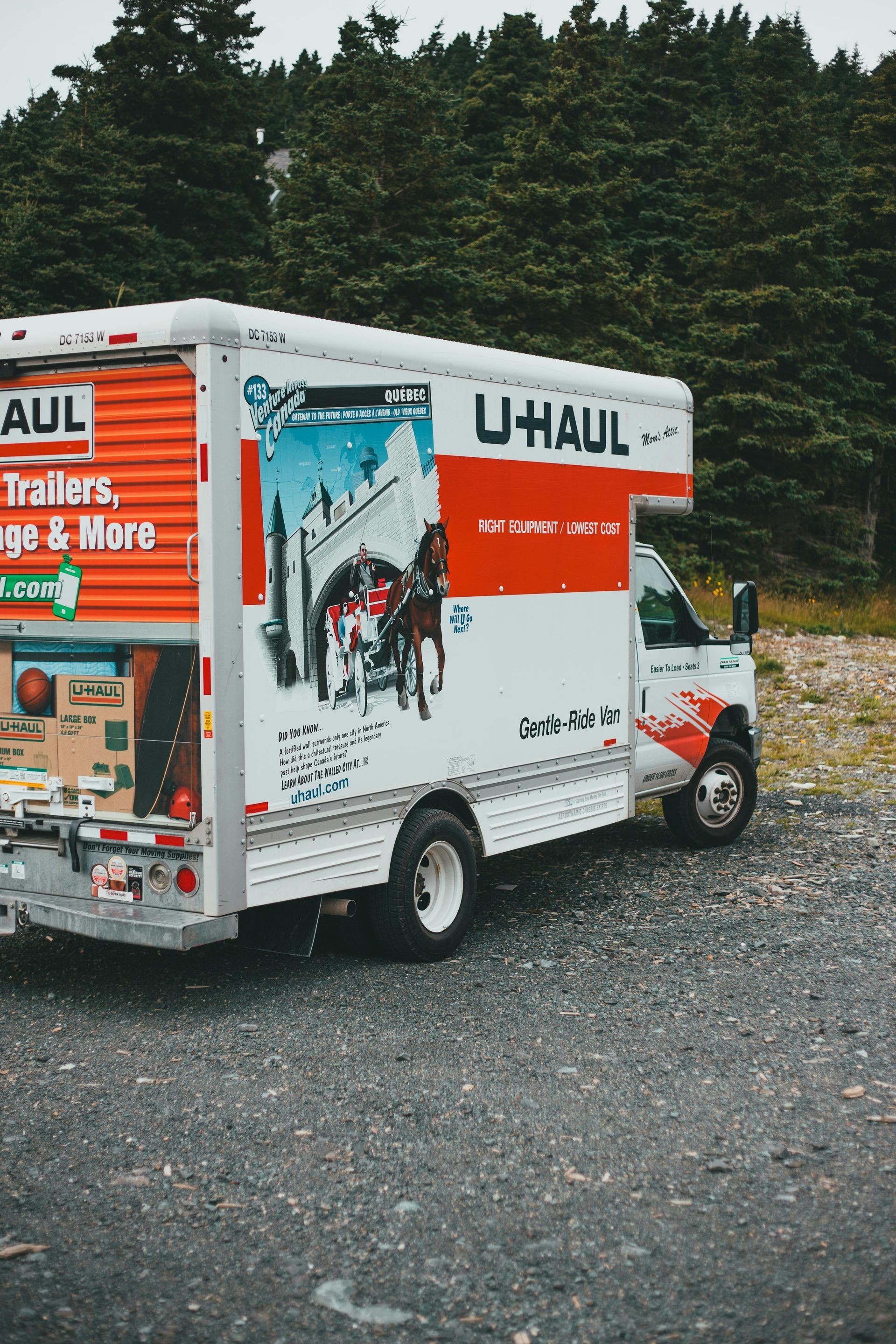 U-Haul moving truck parked on gravel, back door open, boxes visible. Red, orange, white, and blue colors.