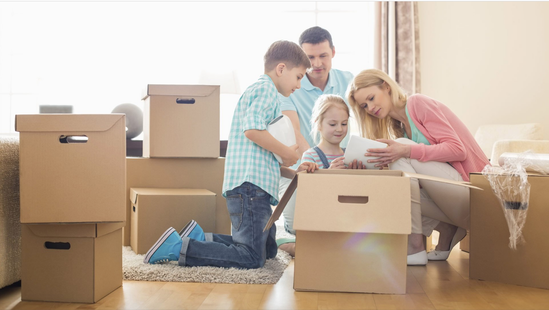 Family packing moving boxes in a bright living room, with laptop and clothes scattered on the floor.