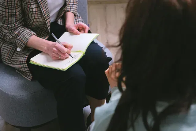 A woman is sitting on a couch talking to another woman while writing in a notebook.