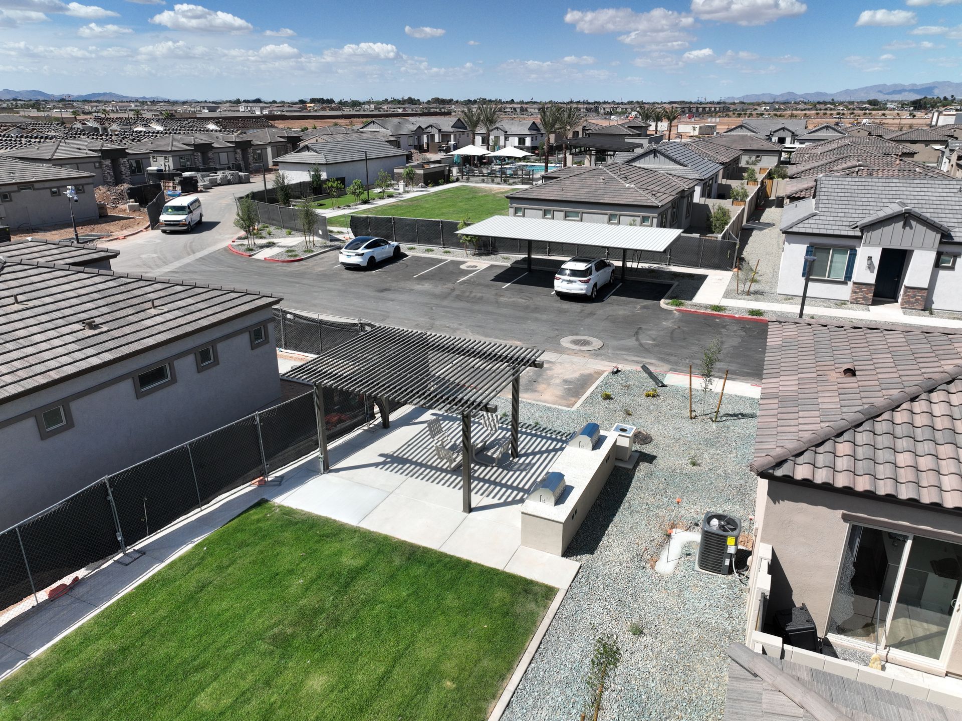 Suburban neighborhood with tiled rooftops, parked vehicles, and a green backyard under a bright blue sky