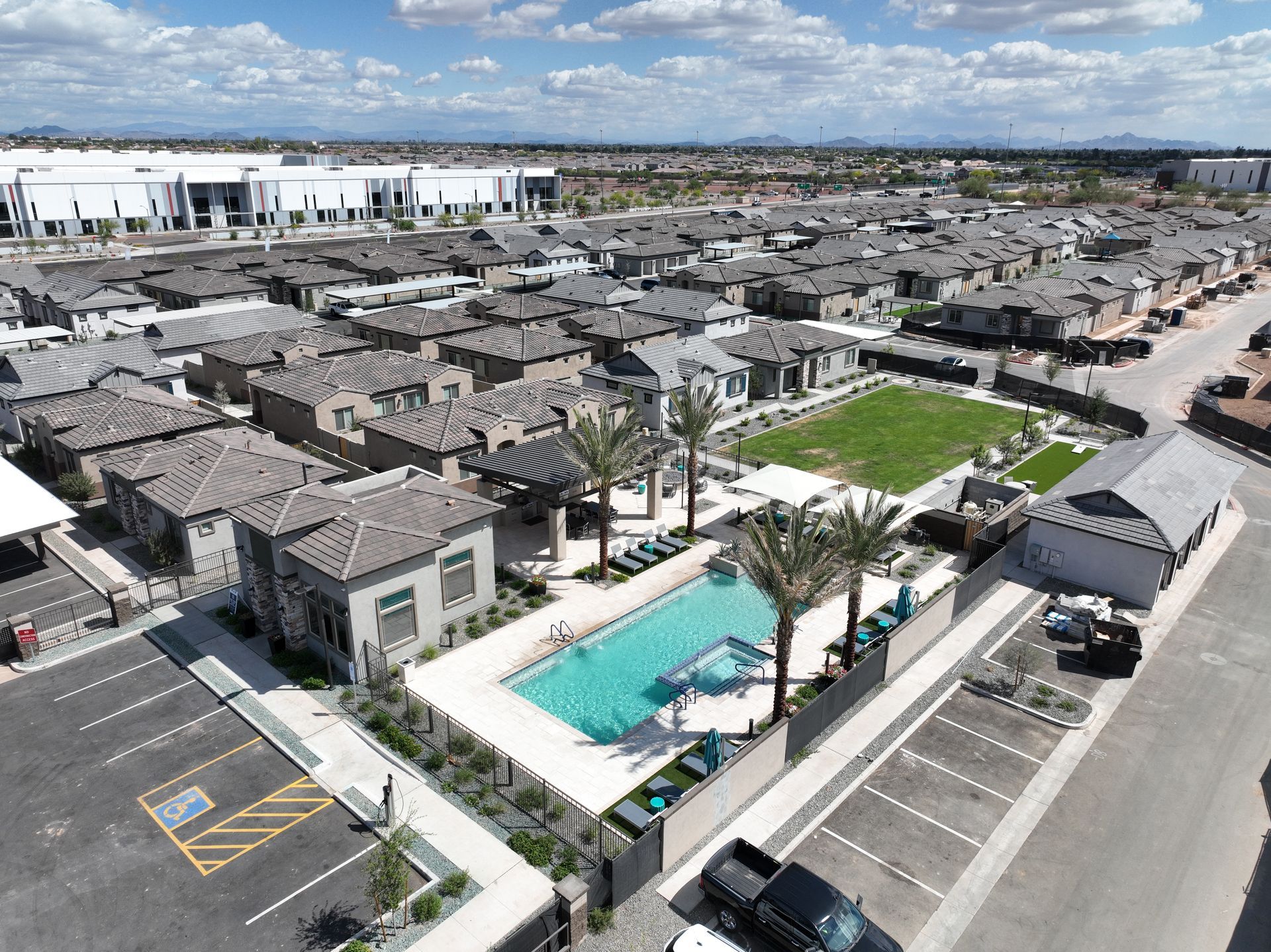 Aerial view of a residential complex with a turquoise pool, palm trees, and parking lots under a partly cloudy sky