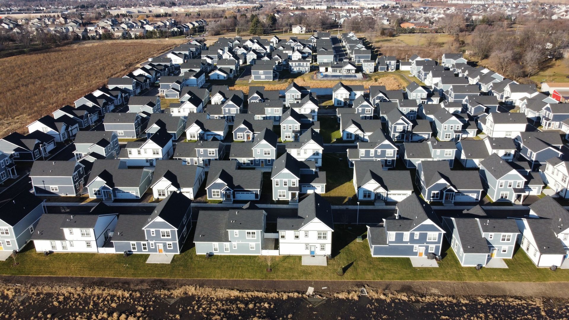 Aerial view of a dense suburban housing development with rows of gray-roofed houses and brown fields nearby