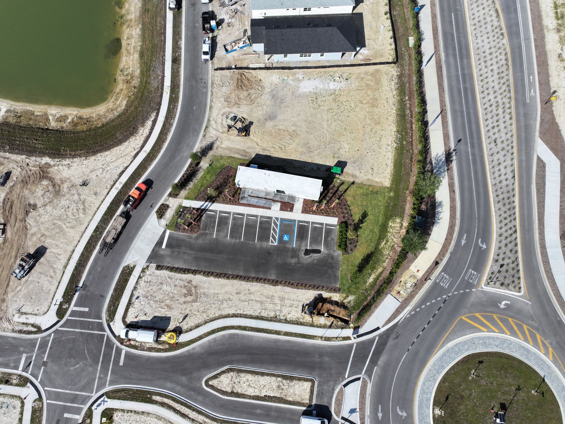 Aerial view of a small building, parked vehicles, and a roundabout beside a waterway.