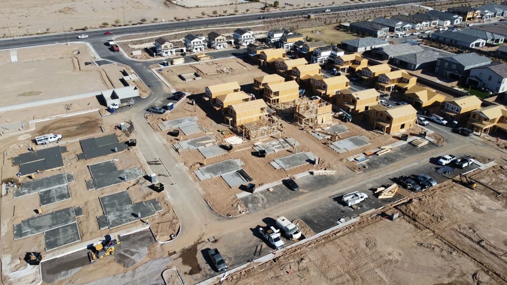 Aerial view of a construction site with rows of tan buildings, dirt roads, parked vehicles, and desert surroundings