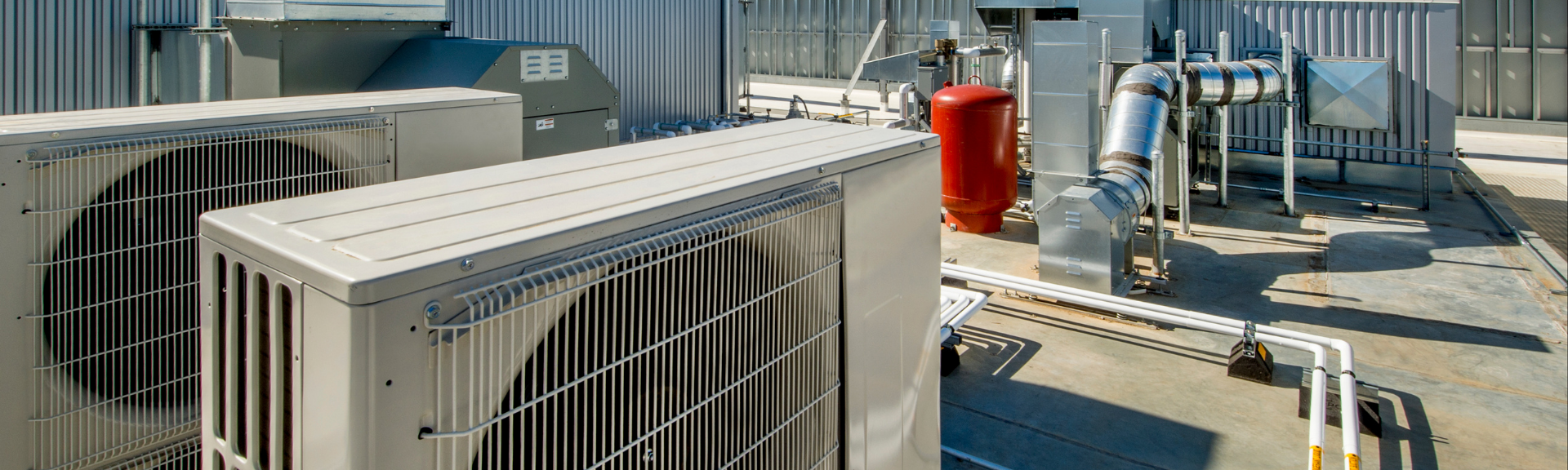 Industrial rooftop HVAC units and piping in a utility area, with metal equipment and a red cylinder.