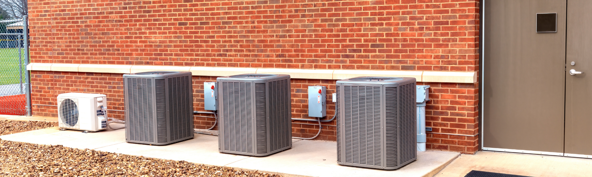 Three outdoor HVAC units against a brick building wall beside a door