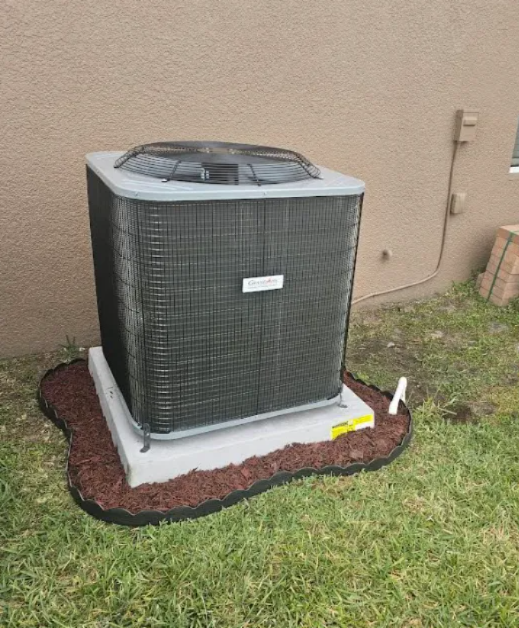 Outdoor central air conditioner unit on a concrete pad beside a beige house, bordered by mulch and grass.