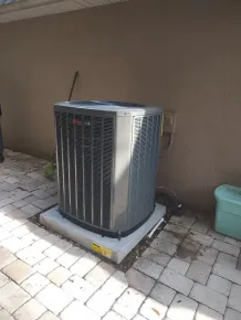 Outdoor air conditioning unit on a patio beside a stucco wall, with brick pavers and a hose nearby.