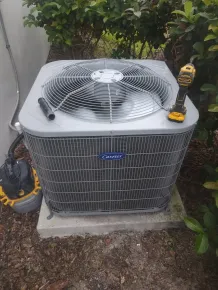 Outdoor central air conditioner unit beside shrubs, with a yellow drill on top and a pressure washer at left.