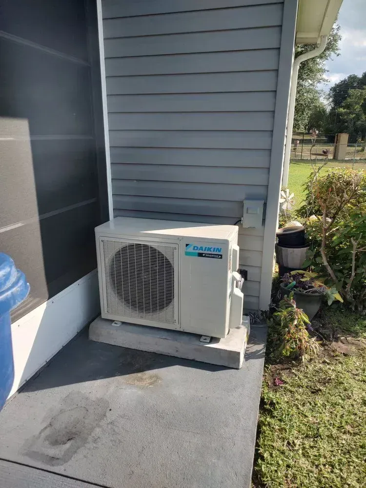 Outdoor air conditioner unit beside a gray house on a concrete pad near a garden.