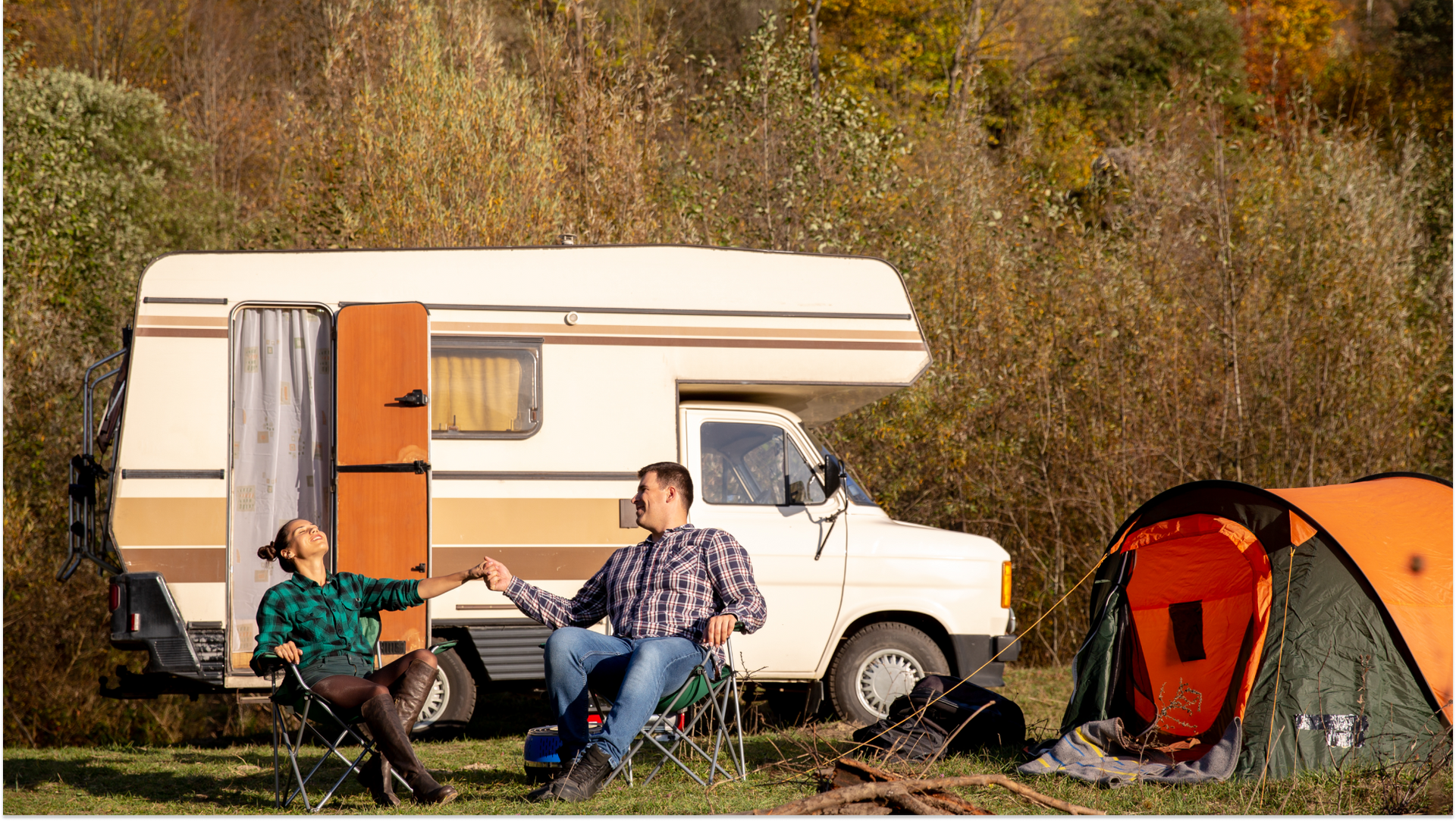 trailer outside and man and woman sitting outside laughing with a camping tent