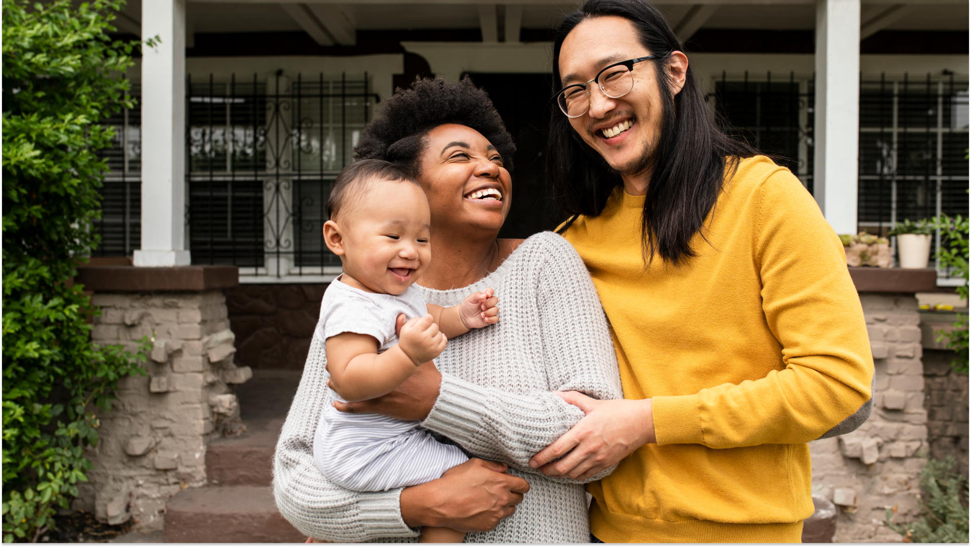 family in various clothing colors smiling with a baby standing outside of a home with green plants