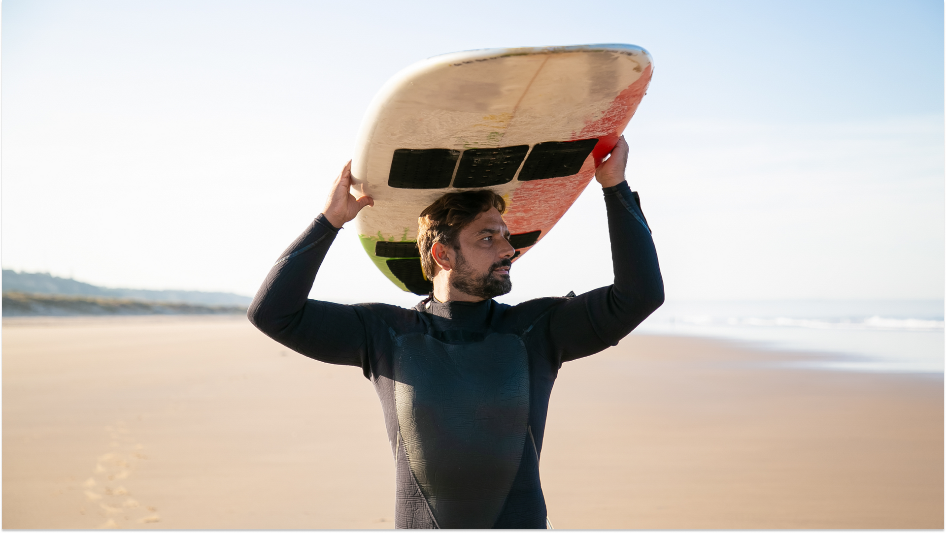 guy on the beach in a wetsuit with a surfboard above his head