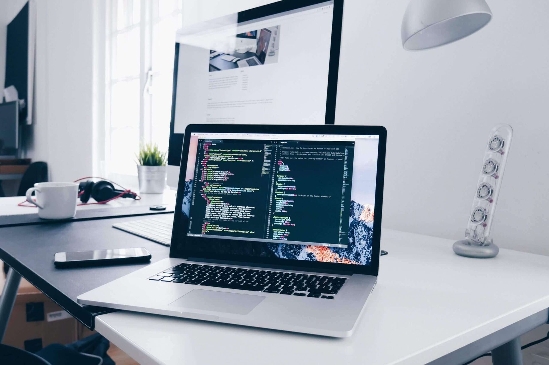 Laptop displaying code on a desk with a monitor, coffee cup, phone, and lamp.