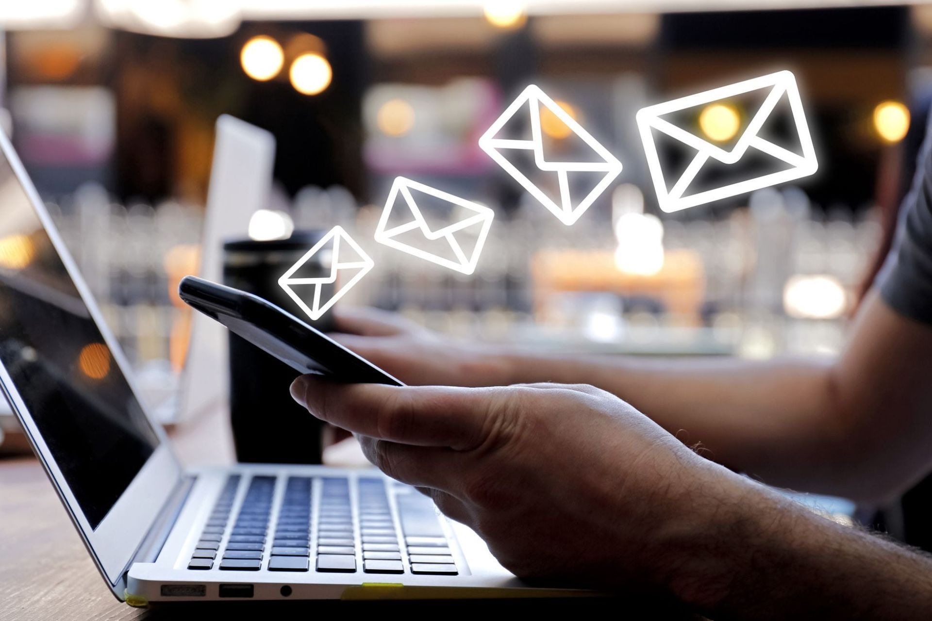 Person holding a phone, sending email, with a laptop in the background. Emails are represented as glowing white icons.