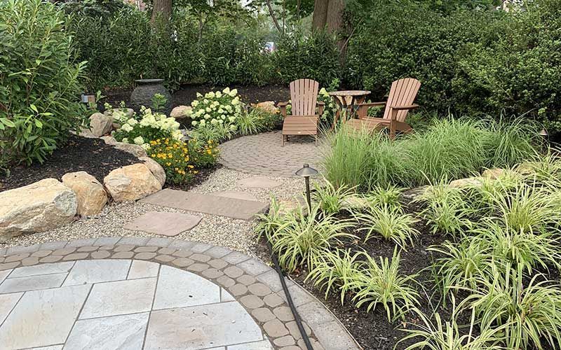 Stone patio with lounge chairs, surrounded by lush garden beds and landscaping.