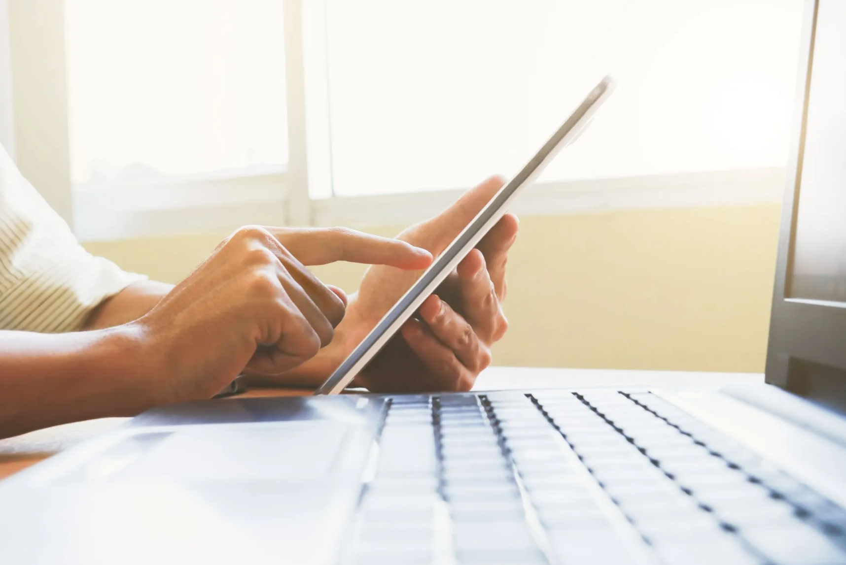 Hands using a tablet over a laptop on a desk; window in background.