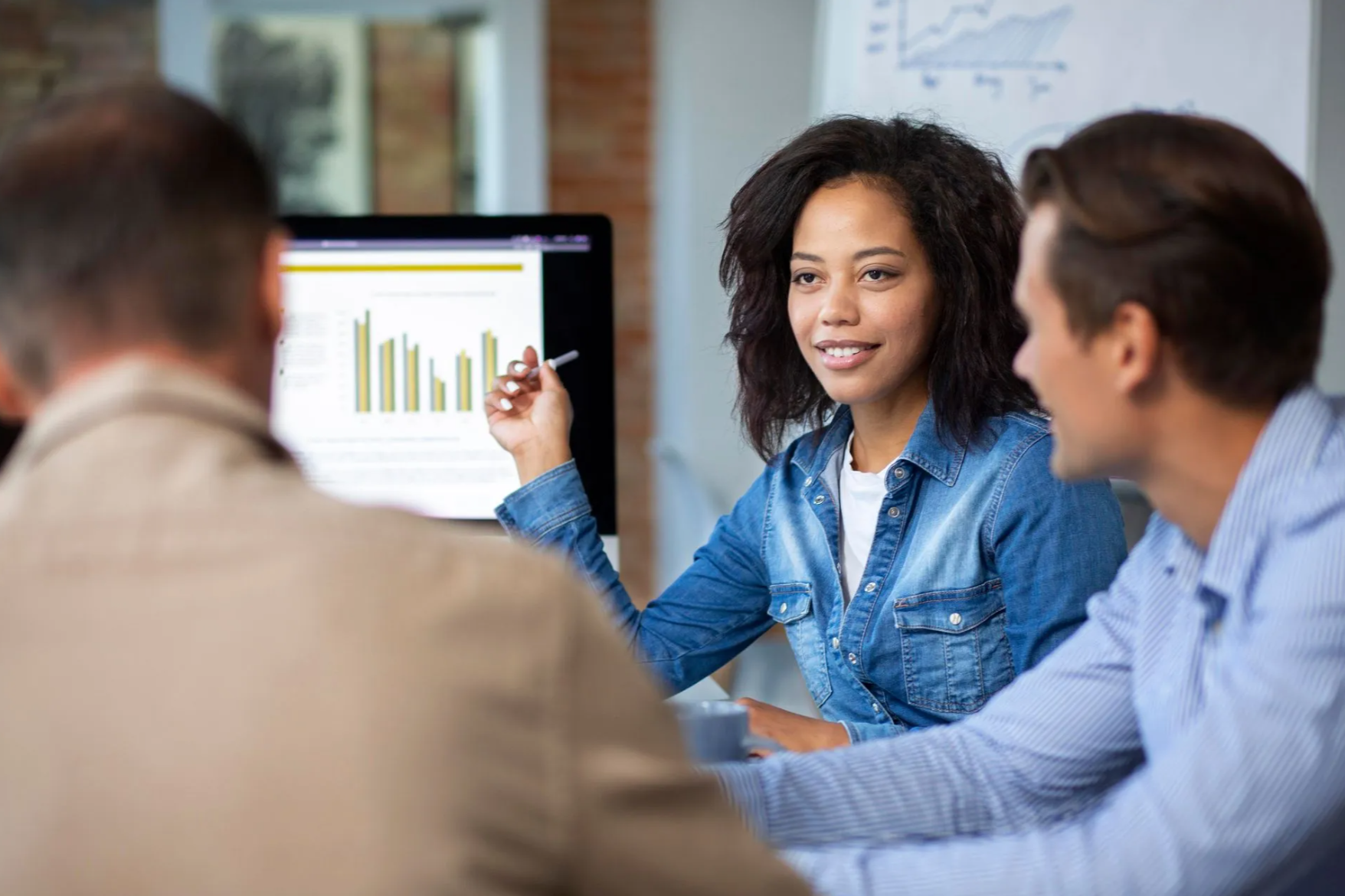Woman pointing at a graph on a computer screen, discussing it with two colleagues in an office.