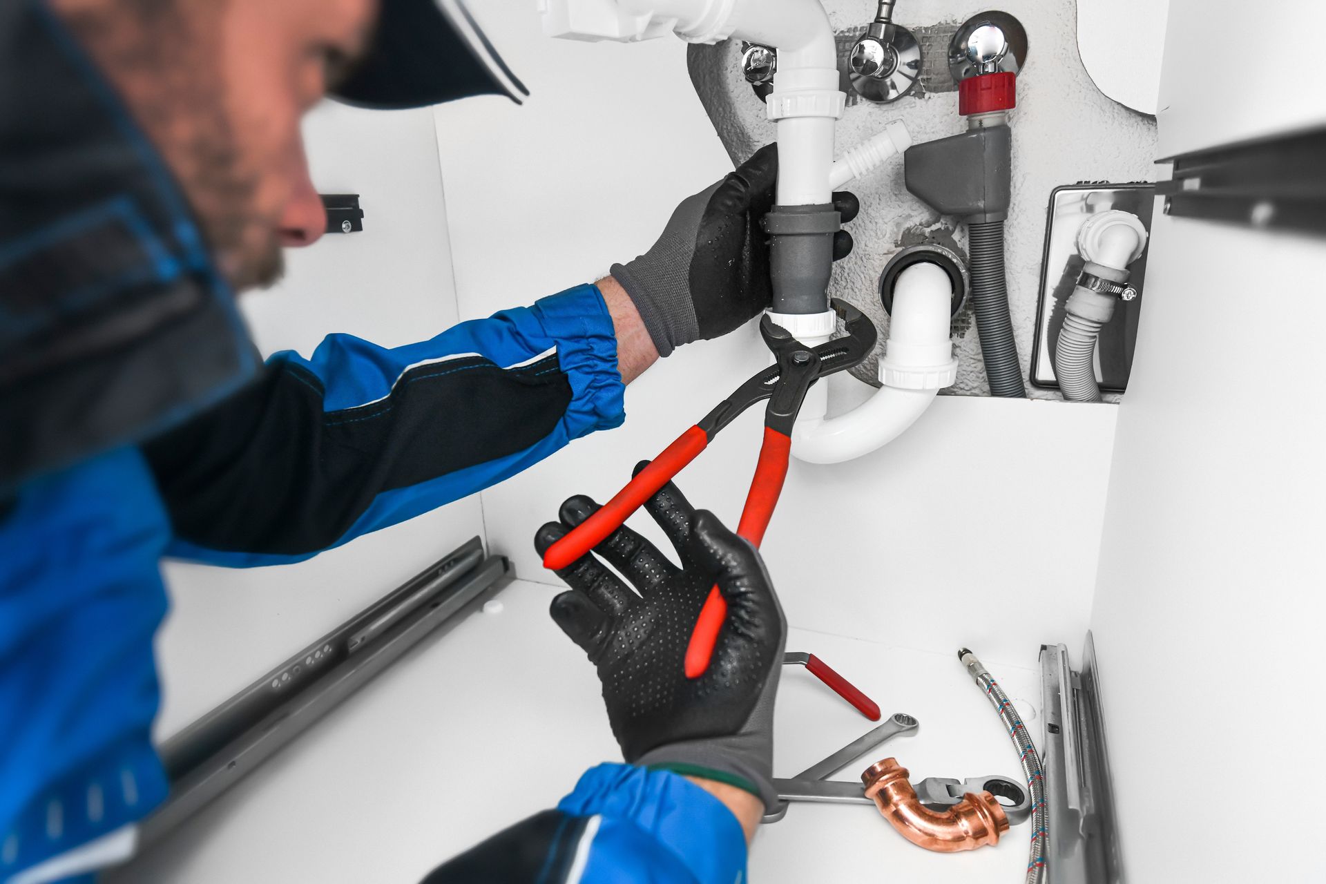 Plumber using pliers on pipes under a white sink, wearing gloves and a blue uniform.