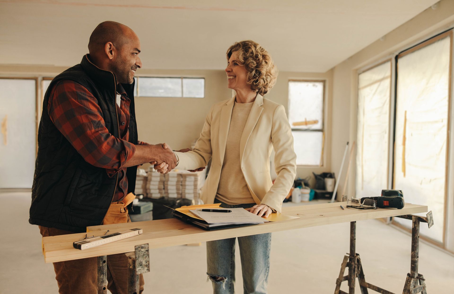 Man in work clothes shakes hands with a woman in a blazer in a construction setting; they smile.