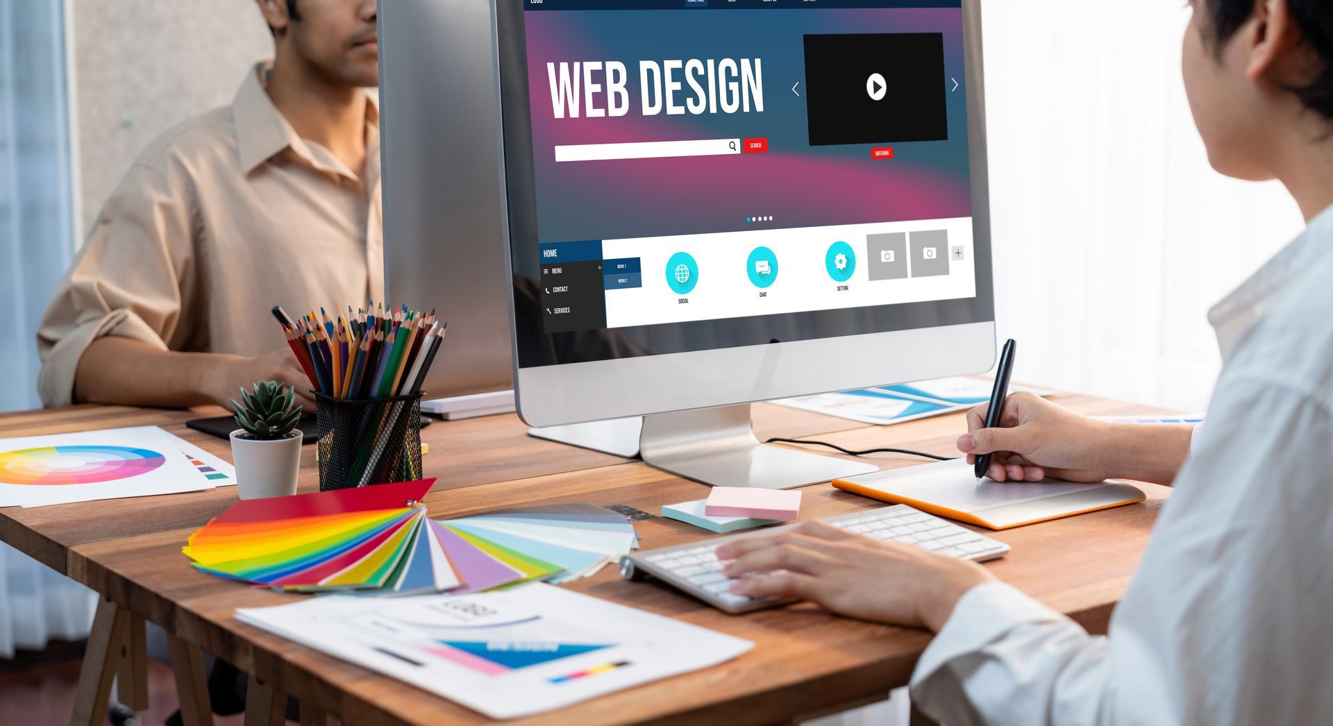 Two people designing a website, using a computer and color swatches on a desk.