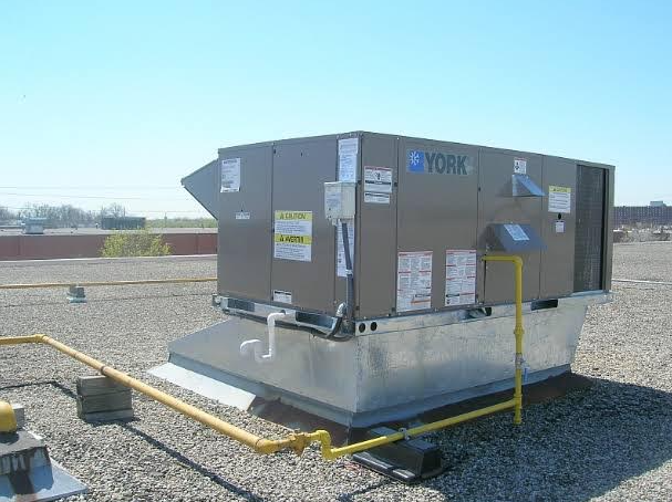 Large gray HVAC unit on a concrete pad in a gravel lot, with yellow piping and open sky.