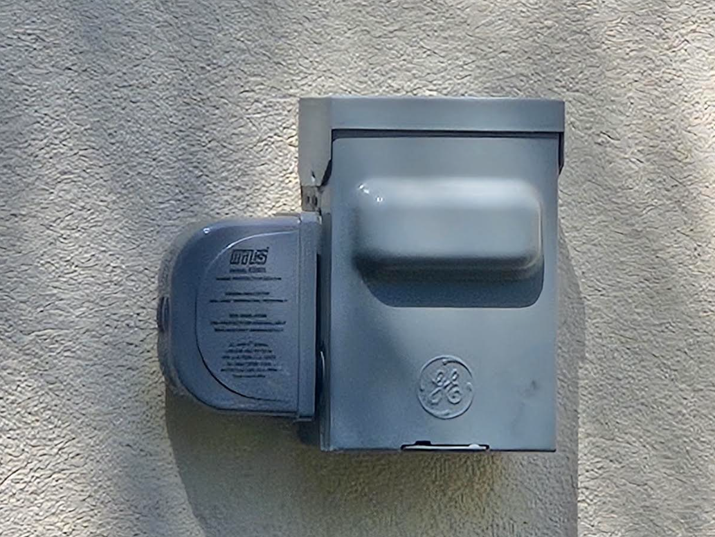 Gray outdoor wall-mounted electrical meter box on a textured stucco wall.