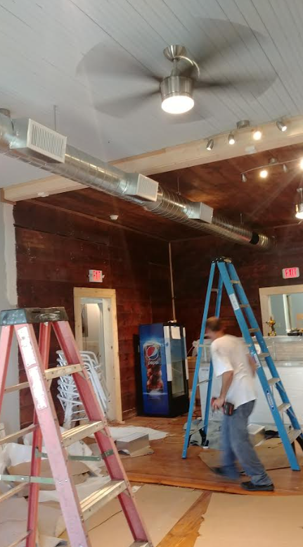 Workers on ladders installing ceiling fixtures in a brick-walled room.