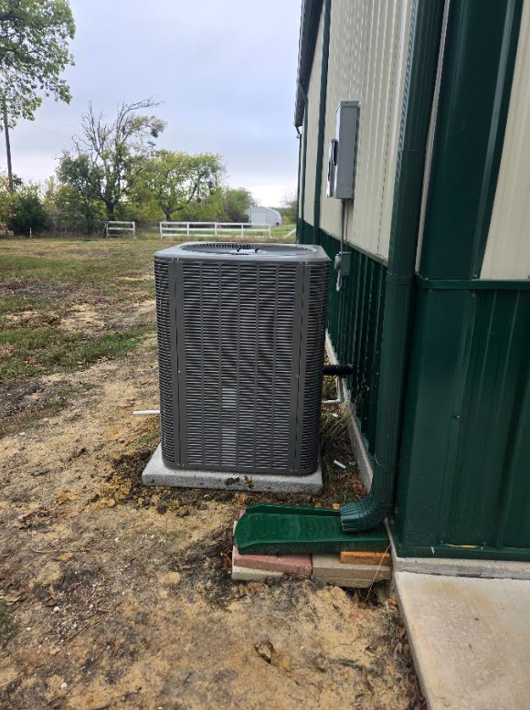 Outdoor air conditioner unit beside a green metal building on a dirt lot