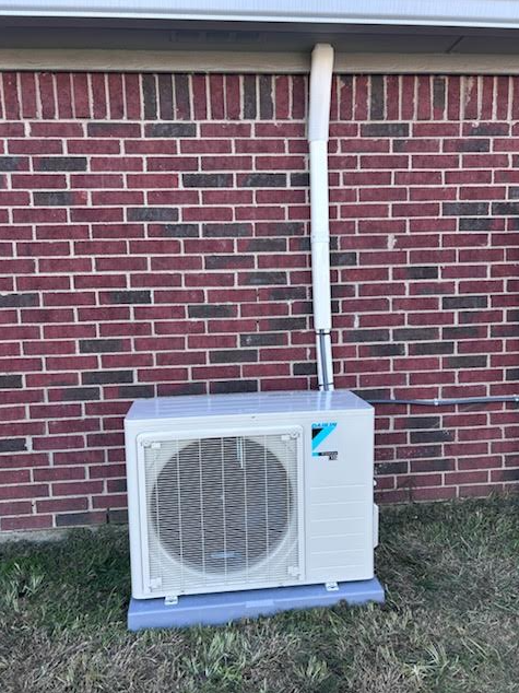 Outdoor AC unit beside a brick wall with exposed piping and wiring