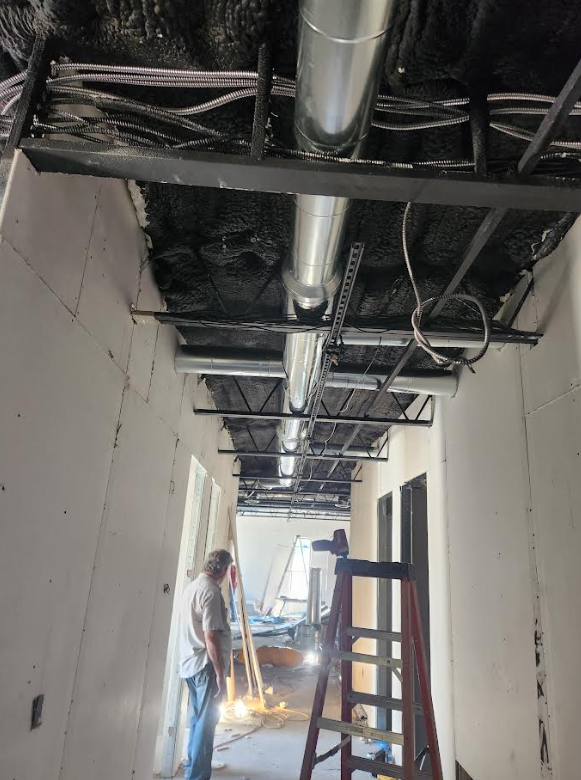 Unfinished hallway with exposed ceiling pipes; worker welding near a ladder and doorway at the far end.