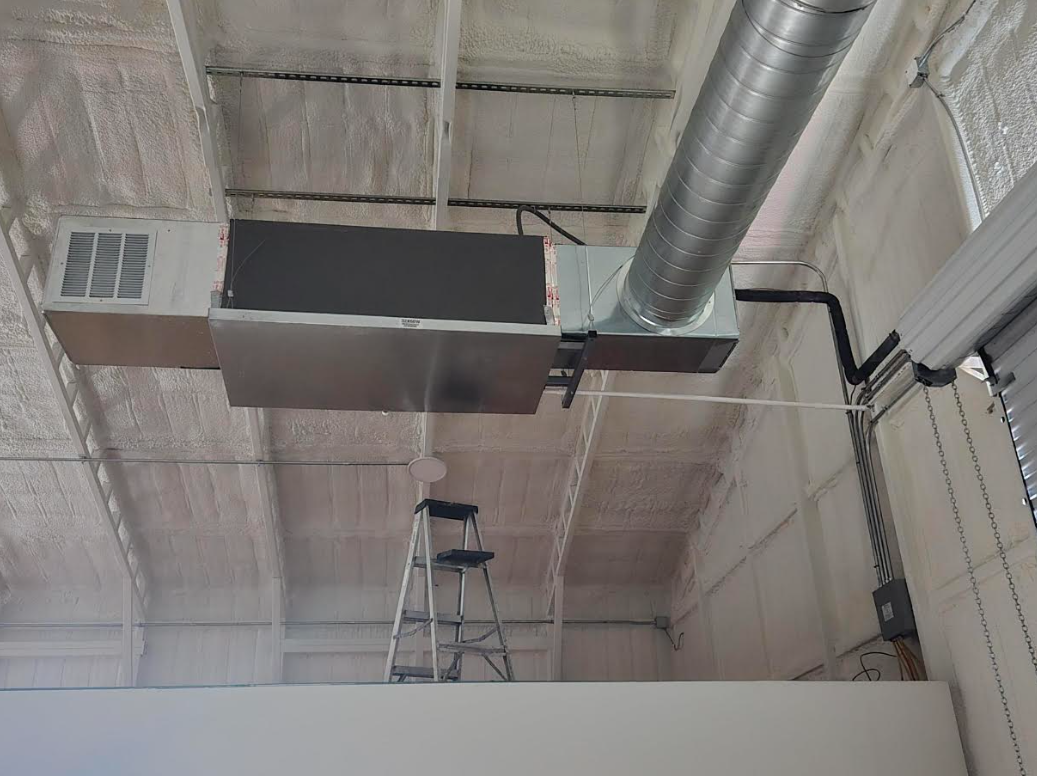 Industrial ceiling with exposed ductwork, a suspended unit, and a ladder below.