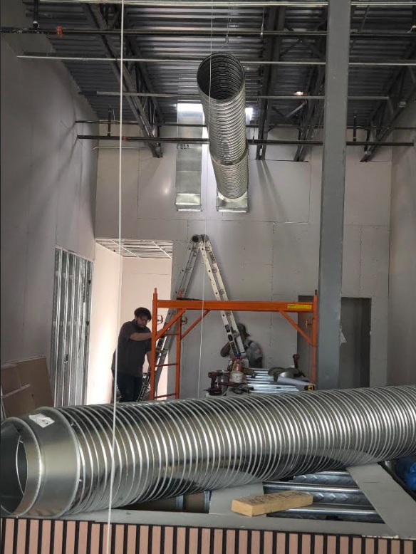 Worker installing large silver ventilation duct in unfinished room with ladder and exposed ceiling