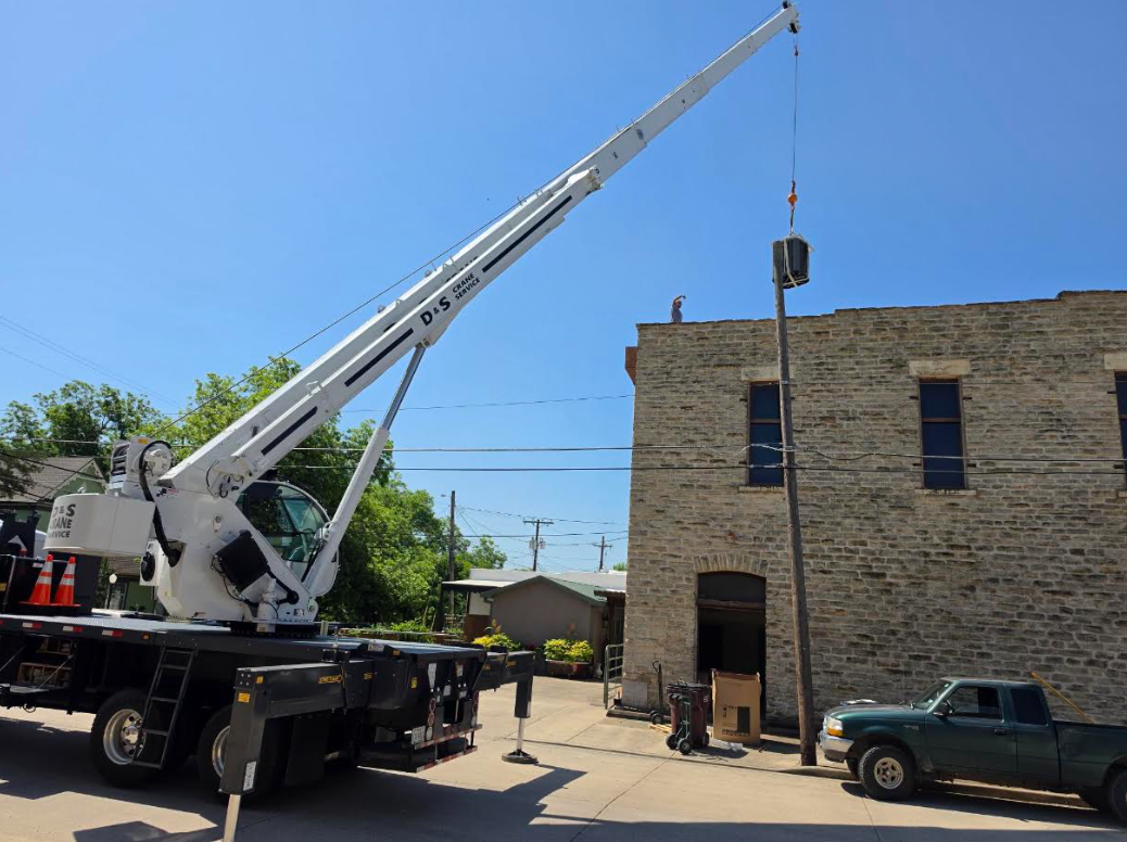 Crane truck lifting a large hanging load beside a brick building on a sunny day