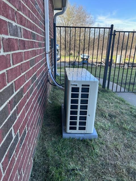 Outdoor air conditioner unit beside a brick house, with a black fence and grassy yard in the background
