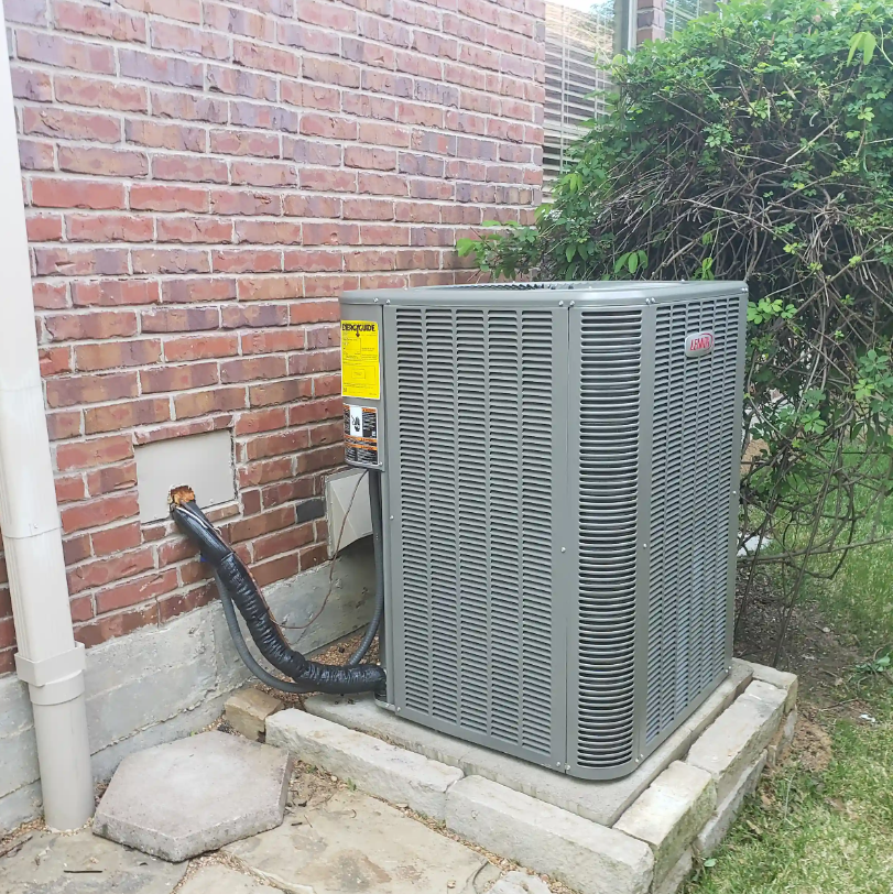Outdoor HVAC unit beside a brick wall, with black insulated piping and shrubs nearby.