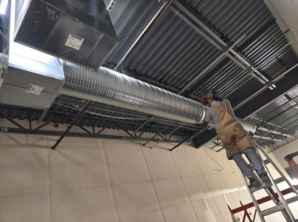 Worker on a ladder installing ductwork in an industrial ceiling with exposed metal beams.