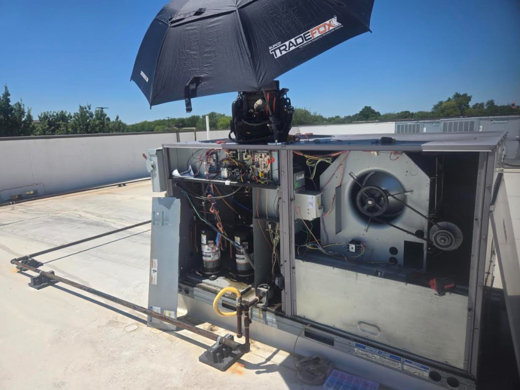 Technician working on an HVAC unit on a rooftop under a black umbrella in bright sun