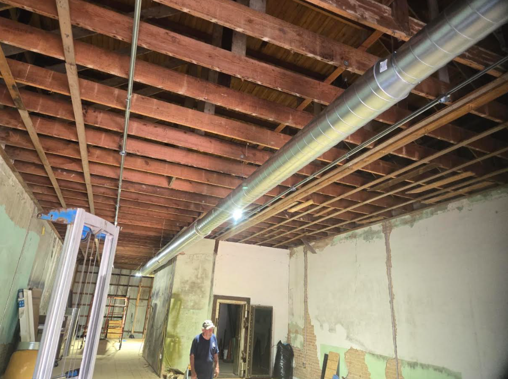 Renovation site with exposed wooden ceiling joists, ductwork, ladder, and a worker below